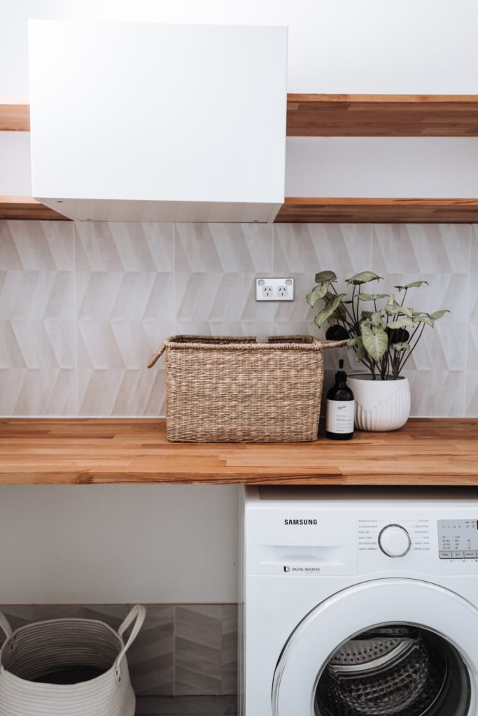 Washing machine sits under a wooden table that supports a plant in a wicker basket.