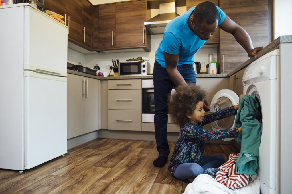 Wide shot of a girl sitting on the floor in the kitchen and helping her father put the washing into the washing machine.